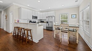 Une cuisine avec des meubles blancs, un îlot central doté de tabourets, une table à manger avec des chaises et un réfrigérateur en acier inoxydable.