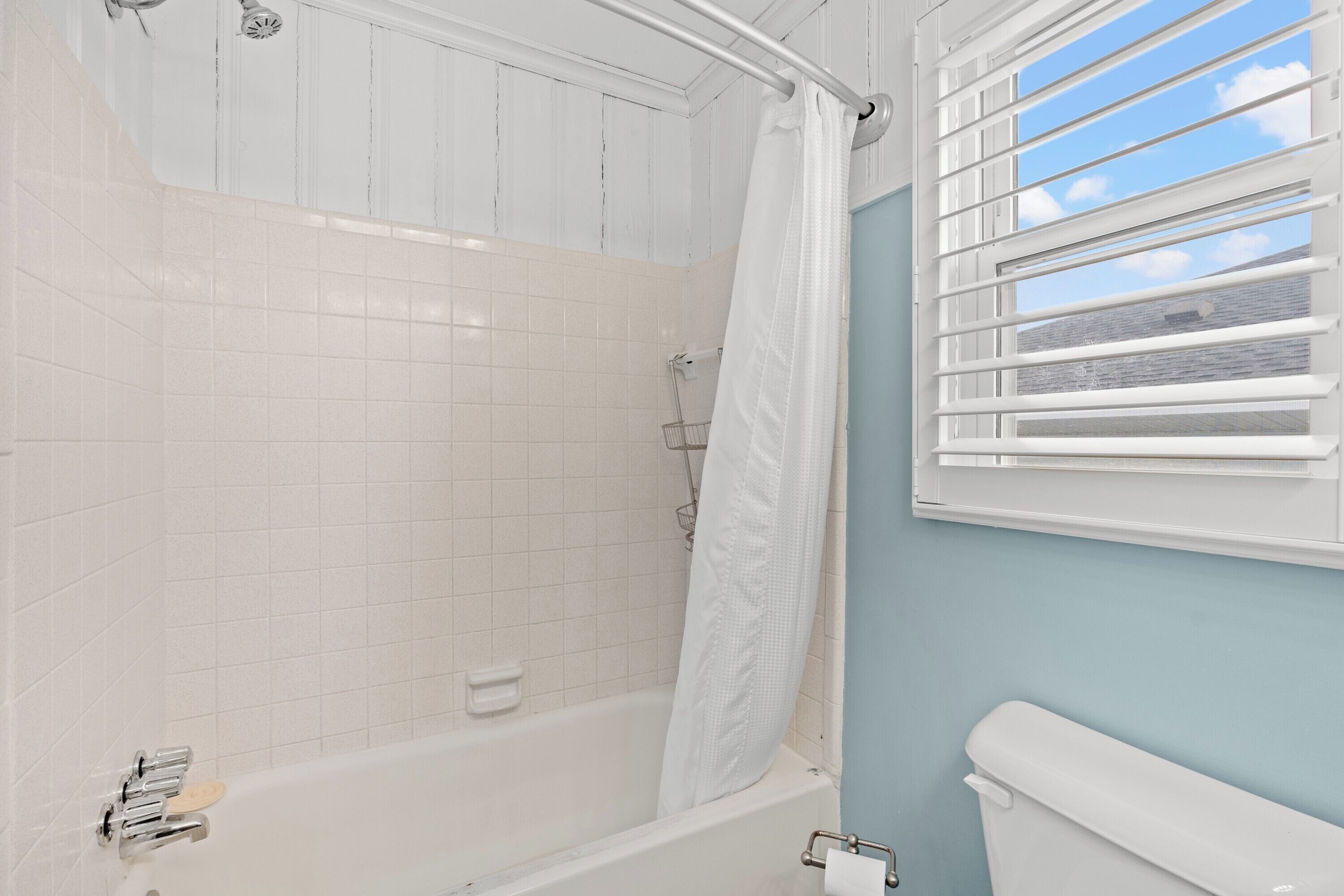 Ein Badezimmer mit Badewanne, Duschvorhang, Toilette und einem Fenster mit Jalousien.