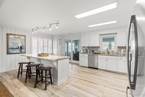 A modern kitchen with white cabinetry, a central island with stools, a refrigerator, and a dining area with a view. - Channel Your Inner Beach, Channel House in Cherry Grove with Fantastic Views (North Myrtle Beach)