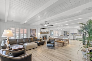 A spacious living area with a sectional sofa, leather armchairs, a wooden coffee table, and a dining table with benches. There is a fireplace, a ceiling fan, and a kitchen in the background. - Channel Your Inner Beach, Channel House in Cherry Grove with Fantastic Views (North Myrtle Beach)