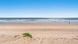 Uma praia com céu claro, areal arenoso e uma pessoa caminhando.