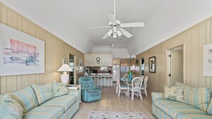A spacious living room with two striped sofas, a dining table with chairs, a ceiling fan, and a kitchen in the background.