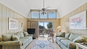 A living room with two sofas, a TV, a ceiling fan, and a large window with a view of palm trees.