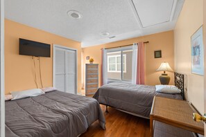 A hotel room with two beds, a TV mounted on the wall, a window with curtains, and a bedside table with a lamp.