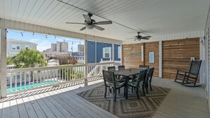 Un espacio al aire libre techado con terraza de madera, una mesa con sillas, ventiladores de techo y vista a una piscina y edificios.
