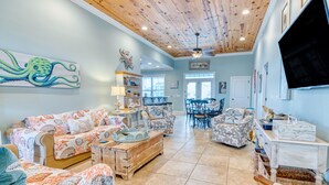 A spacious living room with a high wooden ceiling, a large flat-screen TV, and a dining area with a table and chairs. - Sea La Vie, Luxury Channel Home in Cherry Grove, Beautiful Waterfront Pool Area (North Myrtle Beach)