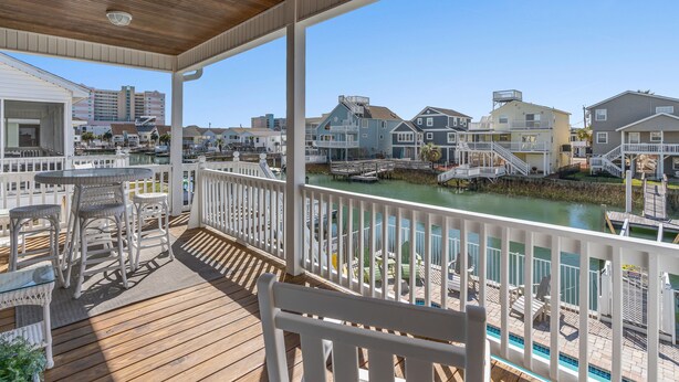A wooden deck with outdoor furniture, a view of a river, and buildings in the background. - Sea La Vie, Luxury Channel Home in Cherry Grove, Beautiful Waterfront Pool Area (North Myrtle Beach)