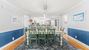 A dining area with a table set for six, surrounded by green chairs, a chandelier, and a blue patterned rug. - Summer Fun - Beach Across the Street (North Myrtle Beach)