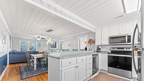 A spacious kitchen with white cabinetry, a central island, stainless steel appliances, and a dining area with a blue sofa and table. - Summer Fun - Beach Across the Street (North Myrtle Beach)