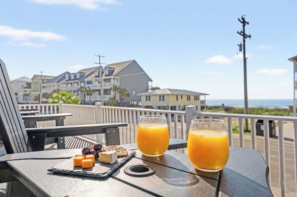 Una mesa con desayuno que incluye queso, galletas y jugo de naranja, con vista a una playa y edificios al fondo.