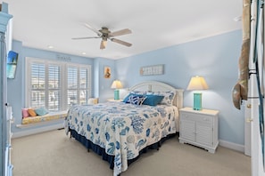 A bedroom with a bed, two bedside tables, a ceiling fan, a window seat, and a door. - Blue Pelican - 5 Bedroom Channel Home - Pet Friendly (North Myrtle Beach)