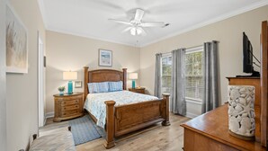 Un dormitorio con cama de madera, dos mesitas de noche con lámparas, un ventilador de techo, una ventana con cortinas y un jarrón decorativo sobre un tocador.