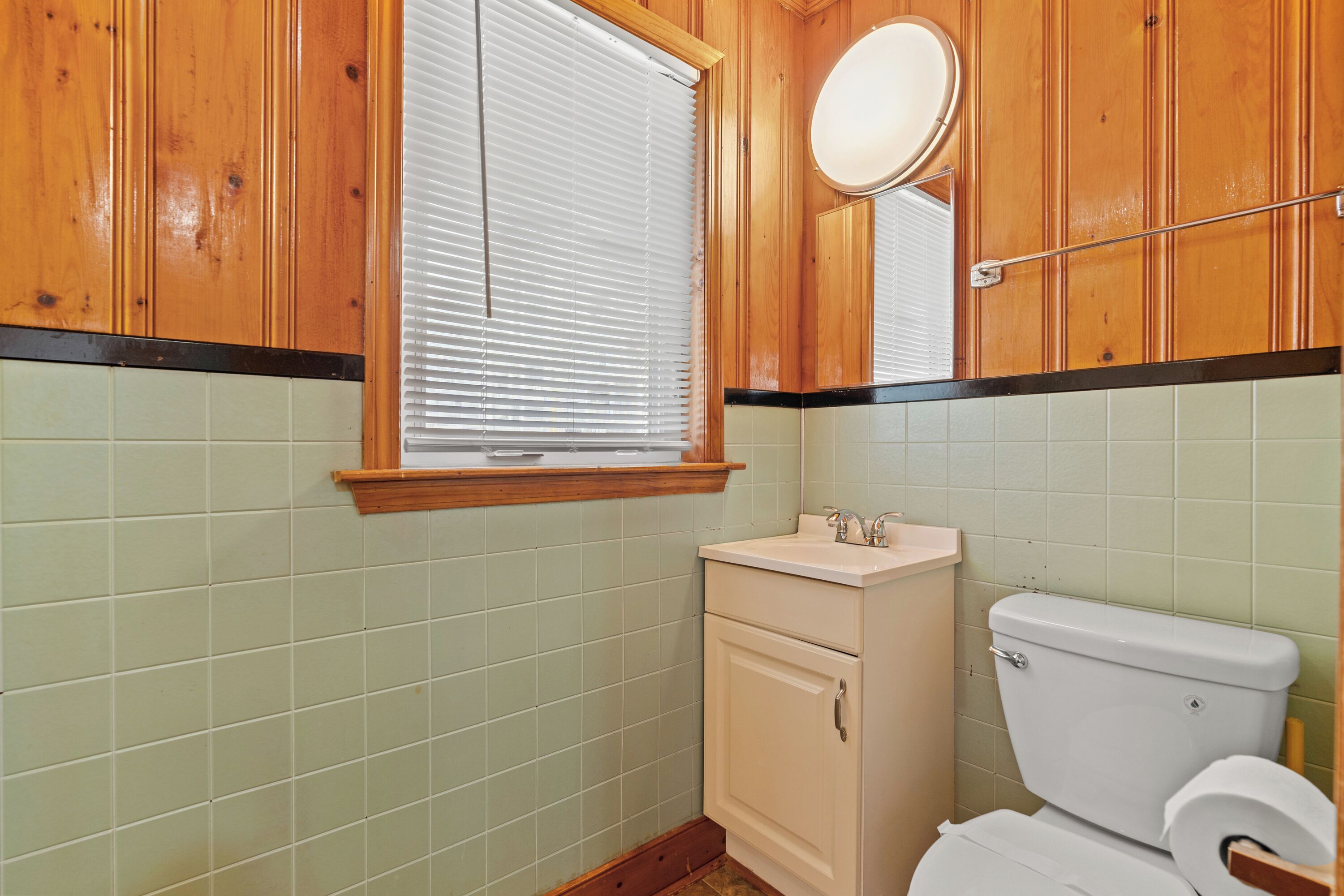A bathroom with wooden cabinets, a white sink, a toilet, and a window with blinds.