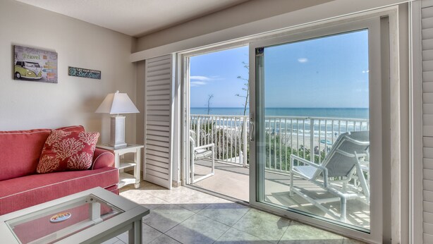 A room with a red sofa, a glass coffee table, a white lamp, and a view of the beach through a sliding glass door.