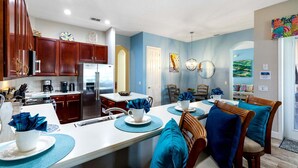 Una cocina moderna con armarios de madera, un refrigerador de acero inoxidable y una zona de comedor con sillas tapizadas en azul y una mesa blanca.