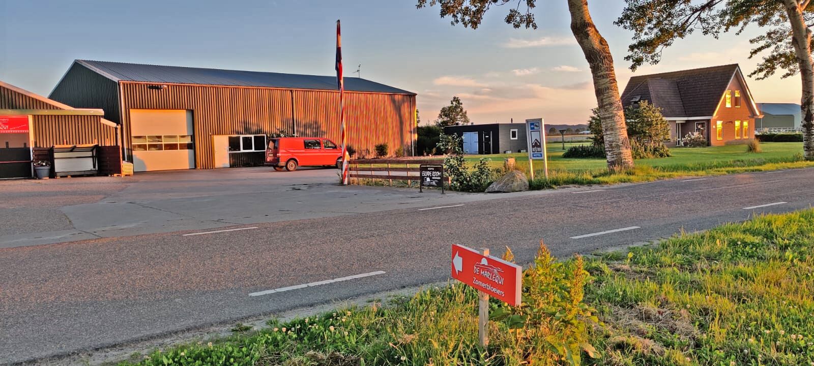 Entorno rural con una furgoneta roja estacionada frente a un edificio que tiene una puerta de garaje grande, una estructura pequeña con el rótulo 'Espace Boom' y un letrero que dice 'De Marequois Zomerbloeiers'.