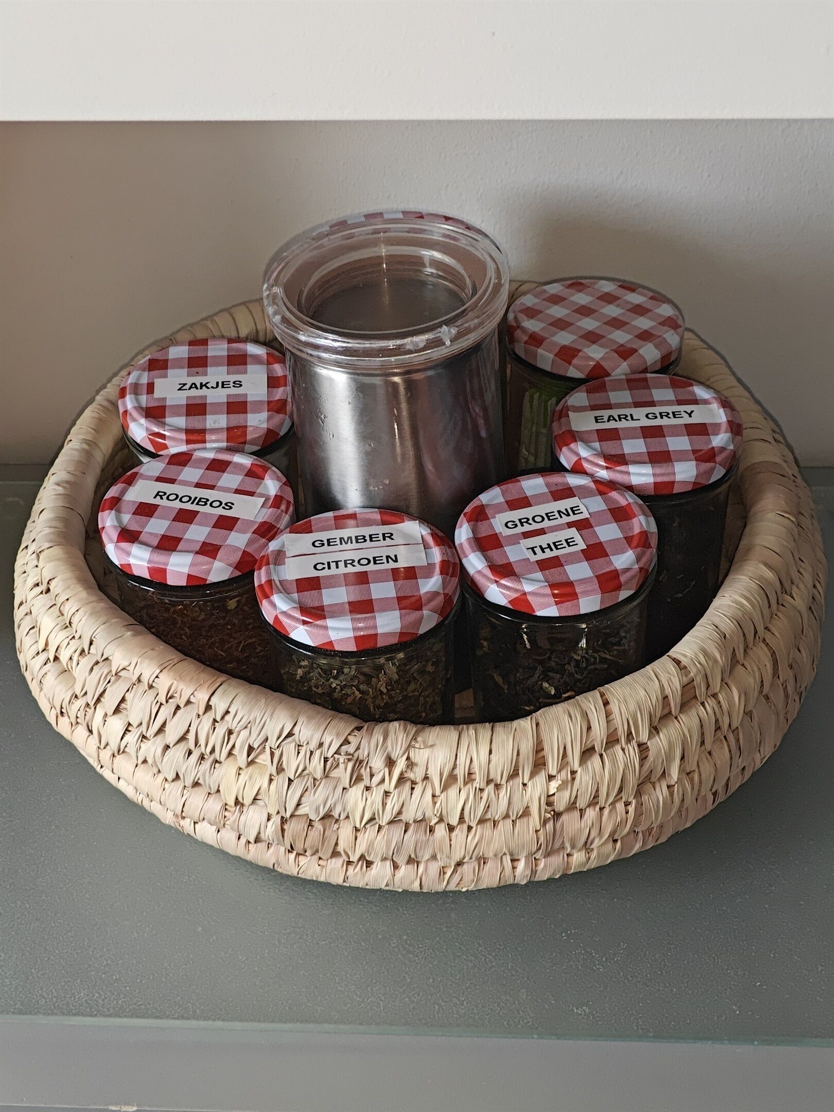 A woven basket containing jars with checkered lids labeled with names like "ZAKJES," "EARL GREY," and "GROENE THEE."