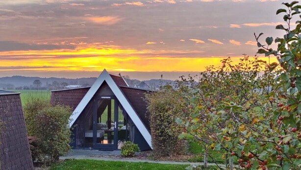 Une maison moderne avec un toit triangulaire et une entrée dotée d’une porte en verre, entourée de verdure et offrant une vue sur le coucher de soleil.