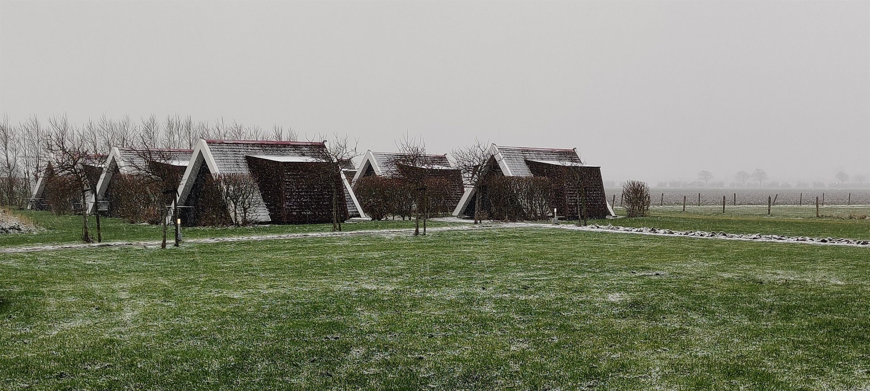 A row of modern, angular buildings with sloped roofs, set in a grassy field with a fence in the background.