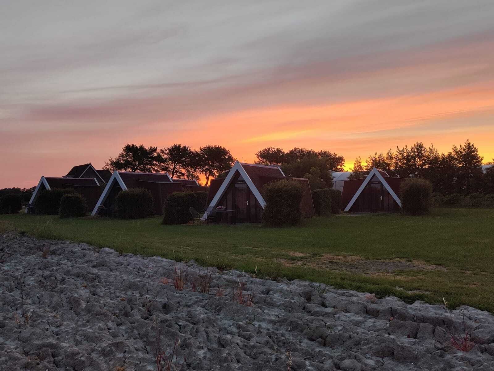 Una hilera de cabañas sencillas con techo a dos aguas, un campo cubierto de césped al frente y un atardecer en el fondo.