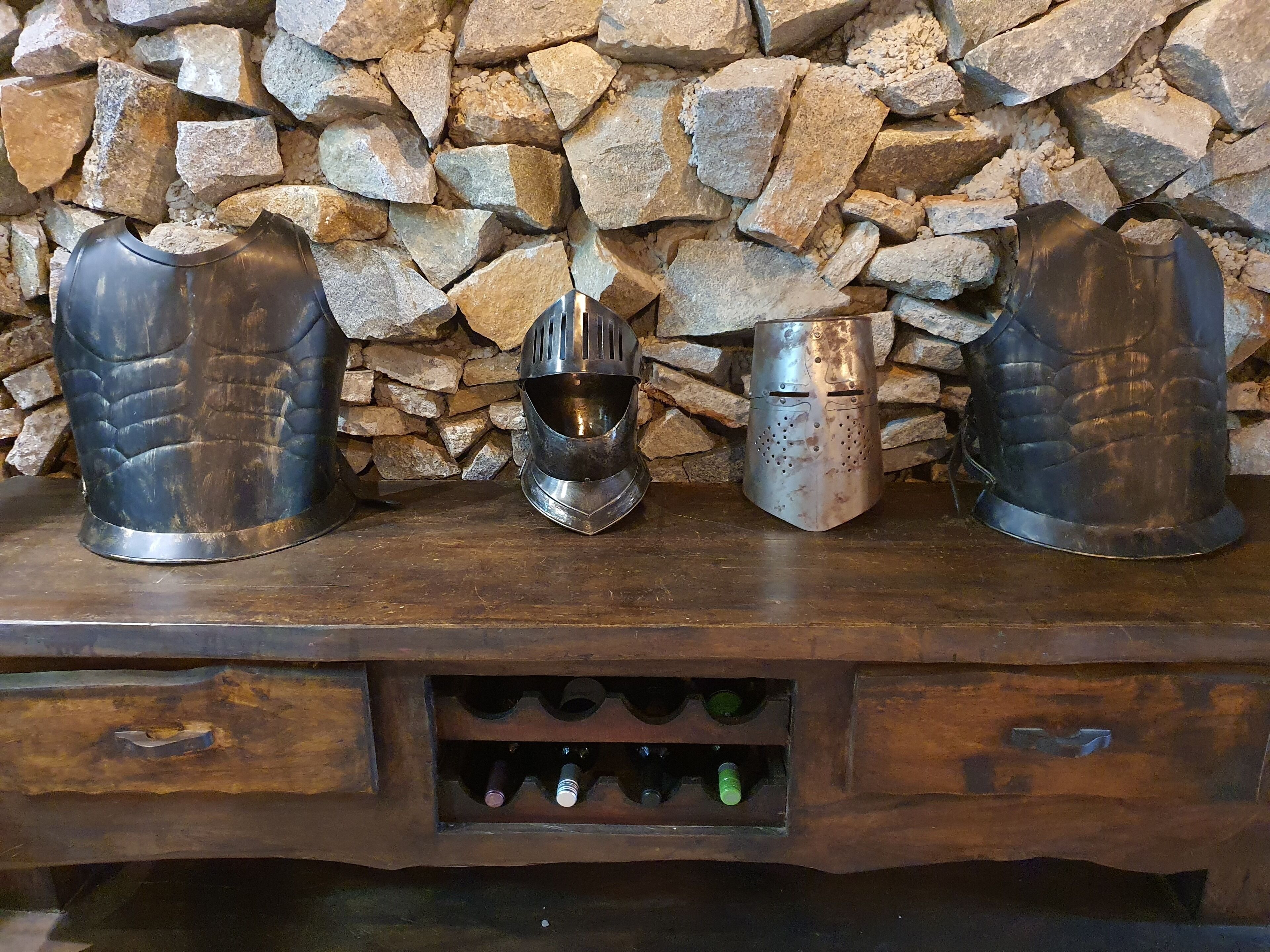 Una mensola di legno con tre oggetti metallici su un muro di pietra. Sotto la mensola, un cassetto di legno con un portabottiglie pieno di bottiglie.