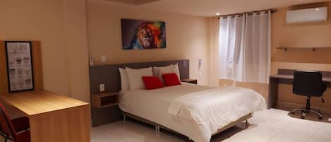 Una habitación de hotel con cama, escritorio, silla, un cuadro y un equipo de aire acondicionado.