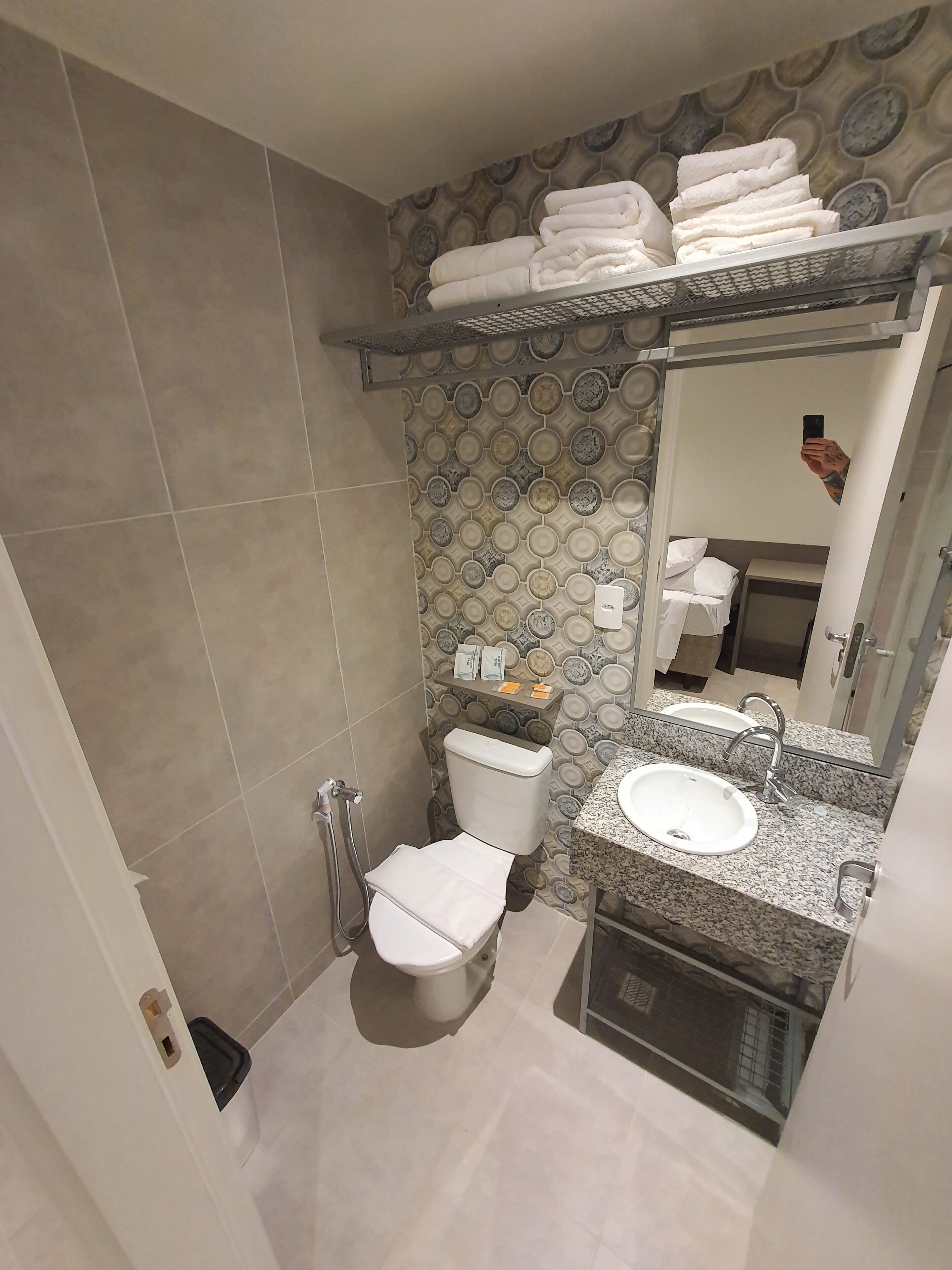 Un baño con inodoro, lavamanos y espejo. Hay un perchero sobre el inodoro y un estante con artículos de aseo.