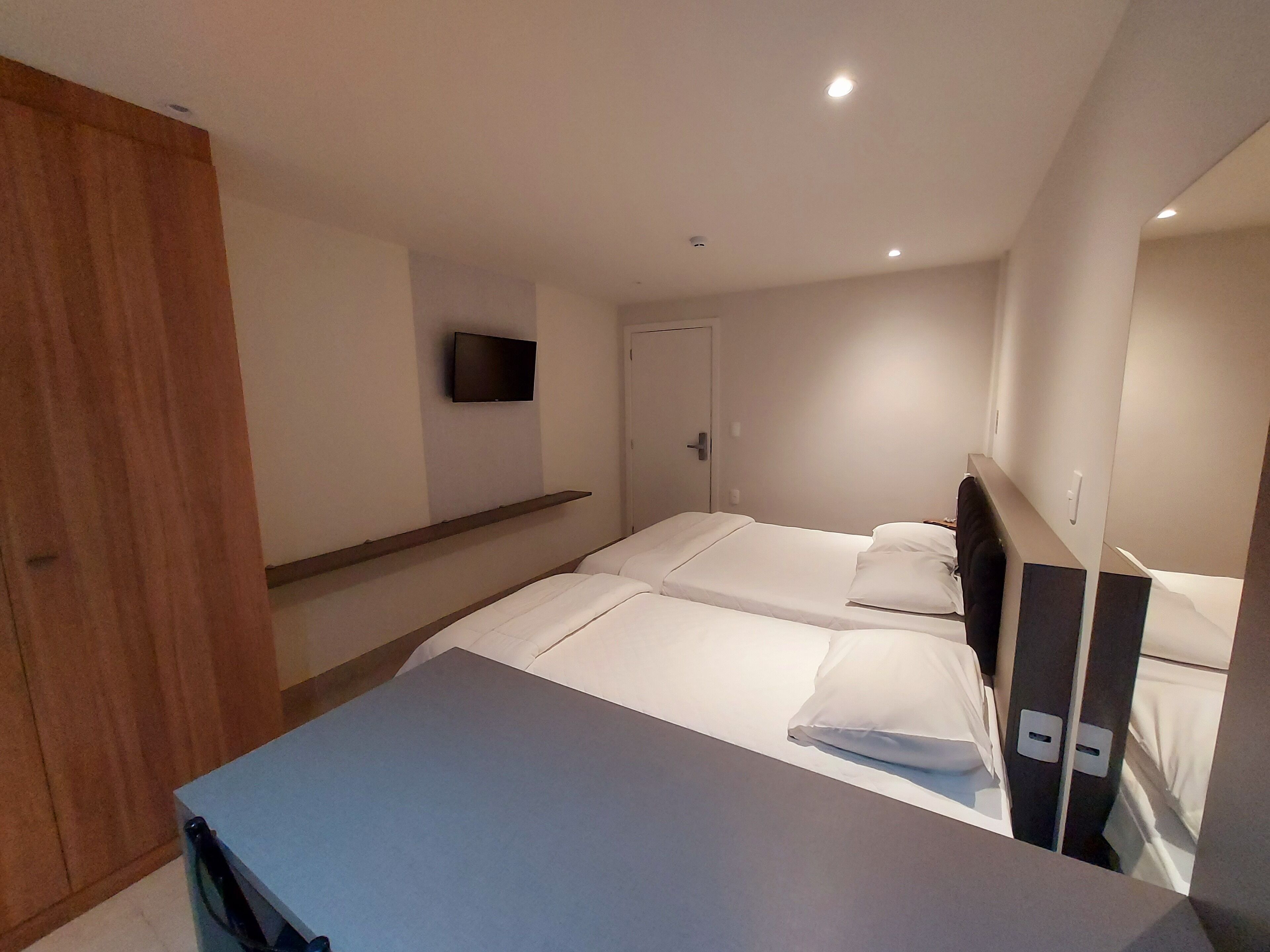 A bedroom with a bed, a TV mounted on the wall, a wooden wardrobe, and a desk.