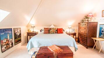 Un dormitorio acogedor con techo inclinado, una cama con manta azul claro, un aparador de madera y una mesita con lámpara.