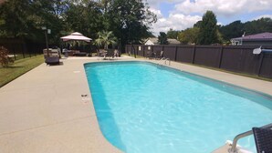A rectangular swimming pool with a metal ladder, surrounded by a concrete deck. There are lounge chairs and a table with umbrellas in the background. - Beaujolais Villas (D'Iberville)