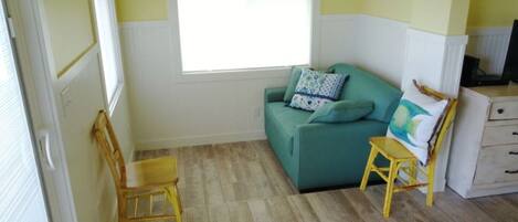 A compact living space with a green sofa, a wooden chair, a small table, and a window providing natural light.