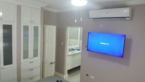 Ett rum med en platt-TV som visar "Smart-TV", en vit garderob med glasdörrar, en vit dörr och en luftkonditioneringsenhet på väggen.