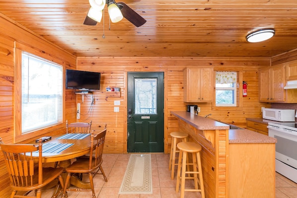 A wooden cabin interior with a dining table, chairs, a kitchen area including a microwave and stove, and a television mounted on the wall.