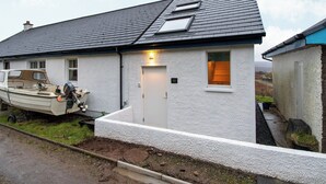 A white house with a grey roof, a boat parked outside, and a small garden area. - 1 bedroom accommodation in Ulva Ferry (Ulva Ferry)