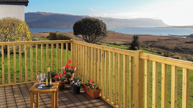 A wooden deck with a table and chairs, potted plants, and a view of a lake and hills. - 1 bedroom accommodation in Ulva Ferry (Ulva Ferry)