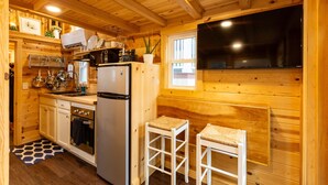 A compact kitchen with wooden walls, a refrigerator, a microwave, and a sink. There are two stools and a window with a view. - Boutique Tiny Home! Pikes Peak Views, HOT TUB, fire pit, & cozy vibes. (Woodland Park)
