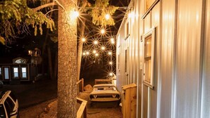 A narrow wooden pathway leading to a building with a metal exterior, surrounded by trees and illuminated by string lights. - Boutique Tiny Home! Pikes Peak Views, HOT TUB, fire pit, & cozy vibes. (Woodland Park)