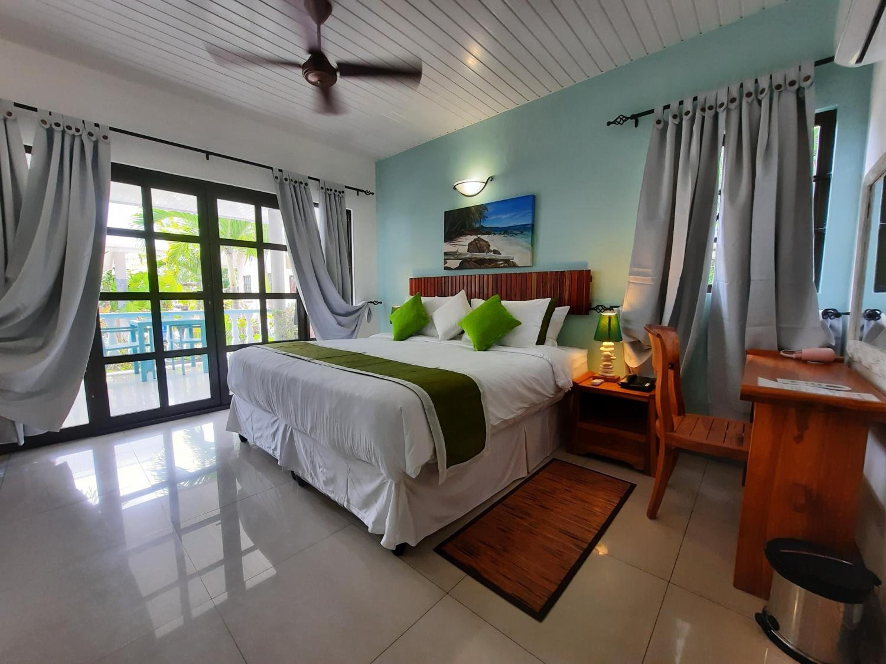 A hotel room with a bed, a desk, a chair, a ceiling fan, and a view of the outdoors.