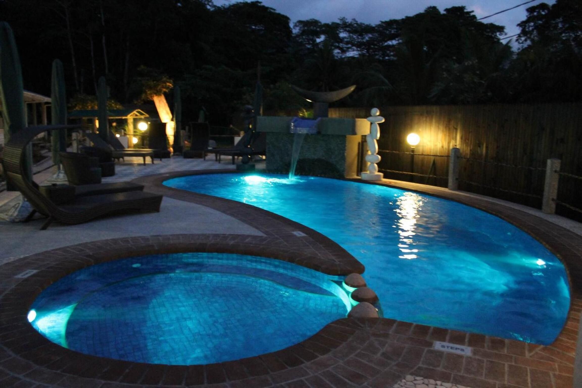 A swimming pool with a waterfall feature, surrounded by lounge chairs and a wooden fence.