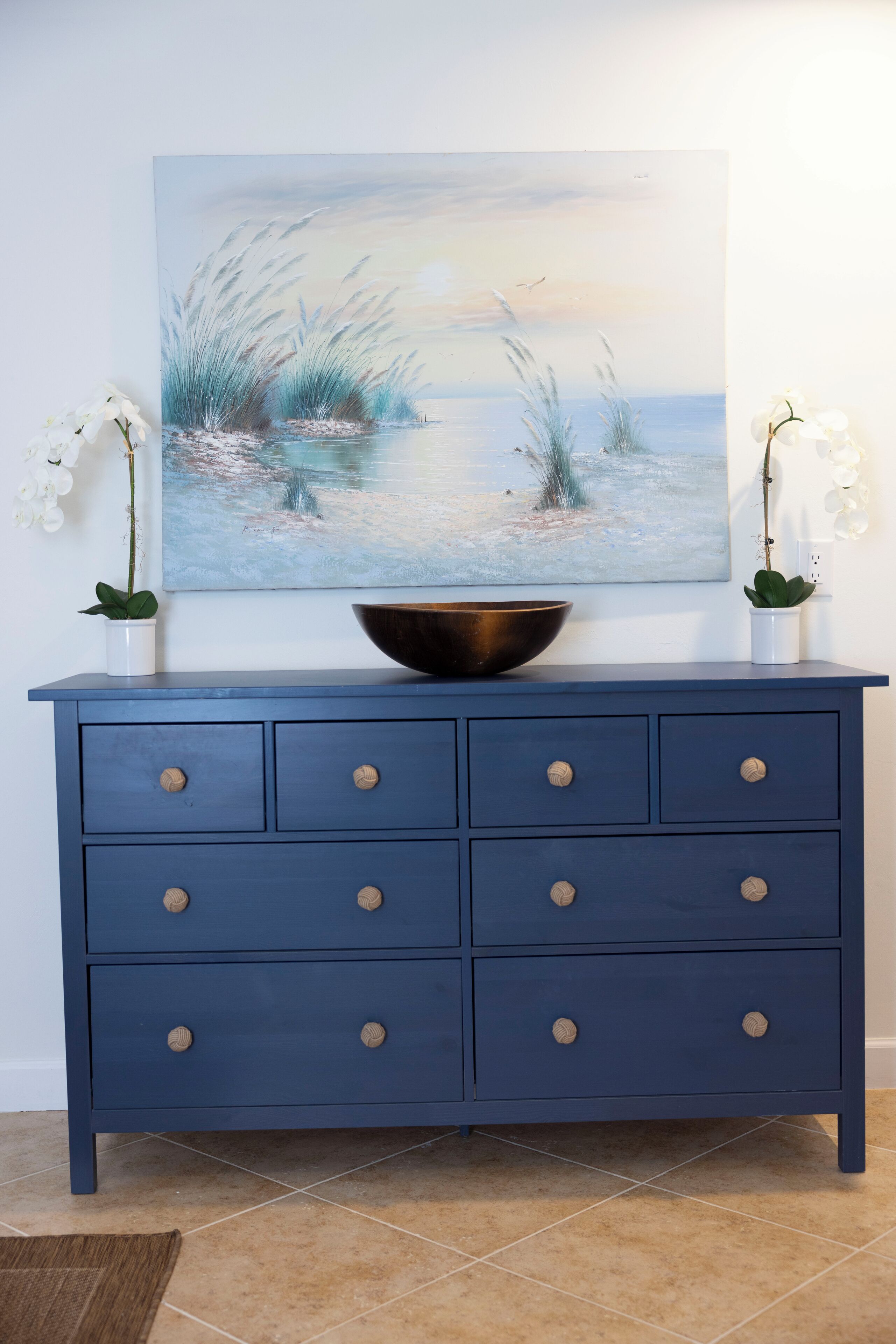 A blue dresser with six drawers, a large painting above it, and two white orchids on either side.