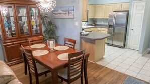 Un coin repas avec une table et des chaises en bois, un meuble avec des portes en verre et une cuisine équipée d’un réfrigérateur et de placards.
