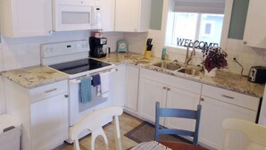 Una cocina compacta con gabinetes blancos, un microondas, una estufa y un lavamanos. Hay una ventana con un cartel que dice 'WELCOME', un reloj pequeño y un florero con flores en la encimera.