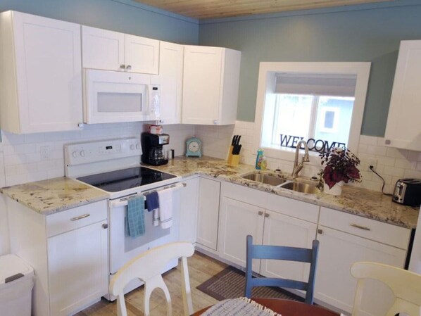 A compact kitchen with white cabinets, a microwave, a stove, and a sink. There's a window with a 'WELCOME' sign, a small clock, and a vase with flowers on the counter.