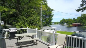 Una terraza con parrilla, mesa de picnic y sillas, con vista a un lago con barcos y casas.