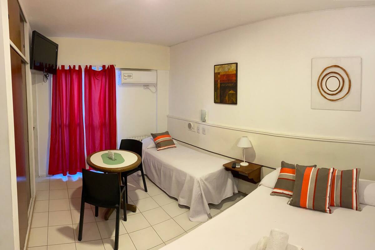 A hotel room with two beds, a small table with a chair, a TV, and an air conditioning unit.