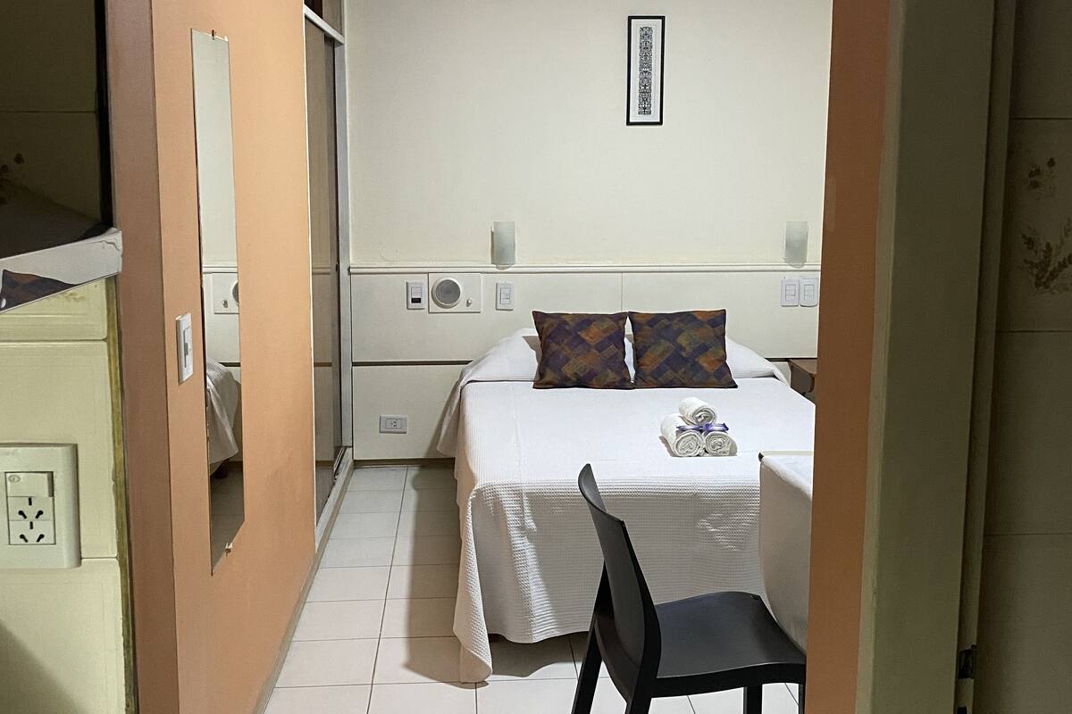 A hotel room with a bed, a chair, a bedside table, a mirror, a wall-mounted light, and a framed picture.