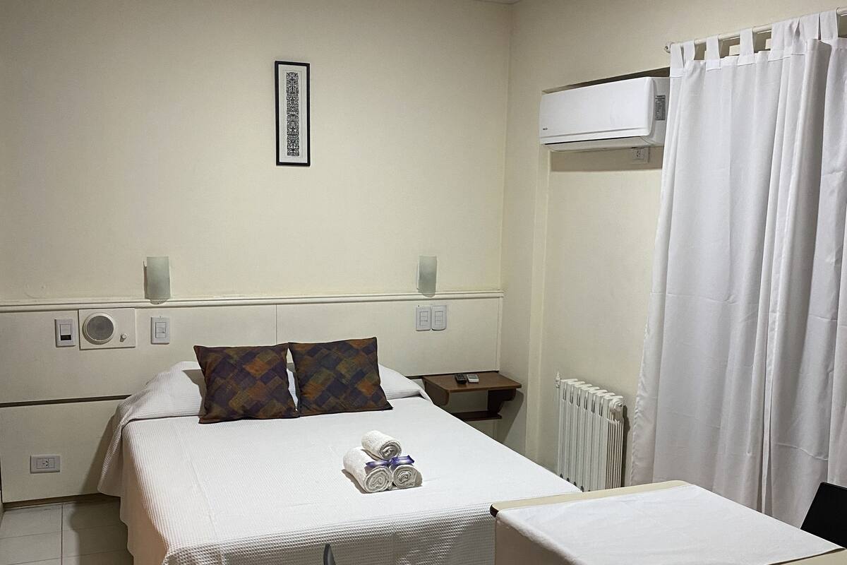 A hotel room with a bed, a table, two chairs, a radiator, and an air conditioning unit.