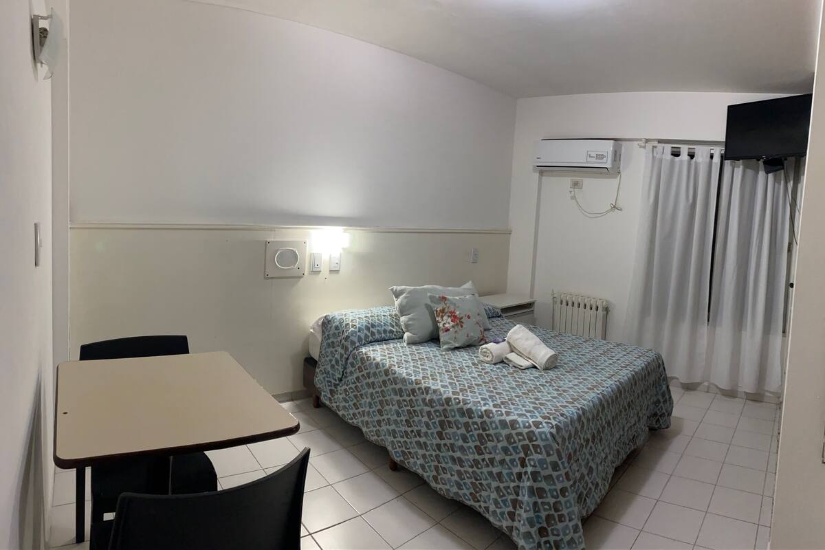 A hotel room with a bed, a desk, a chair, a radiator, and a TV on the wall.