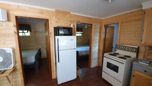 Cocina compacta con refrigerador, microondas, estufa y ventilador. Se observa una cama y una puerta de madera que da a otra habitación.