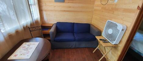 Ein gemütliches Zimmer mit Holzboden, einem blauen Sofa, einem kleinen Tisch mit floralem Untersetzer, einem an der Wand befestigten Fernseher und einem Standventilator.